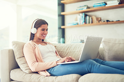 Buy stock photo Shot of a smiling young woman listening to music on a laptop while relaxing on the sofa at home