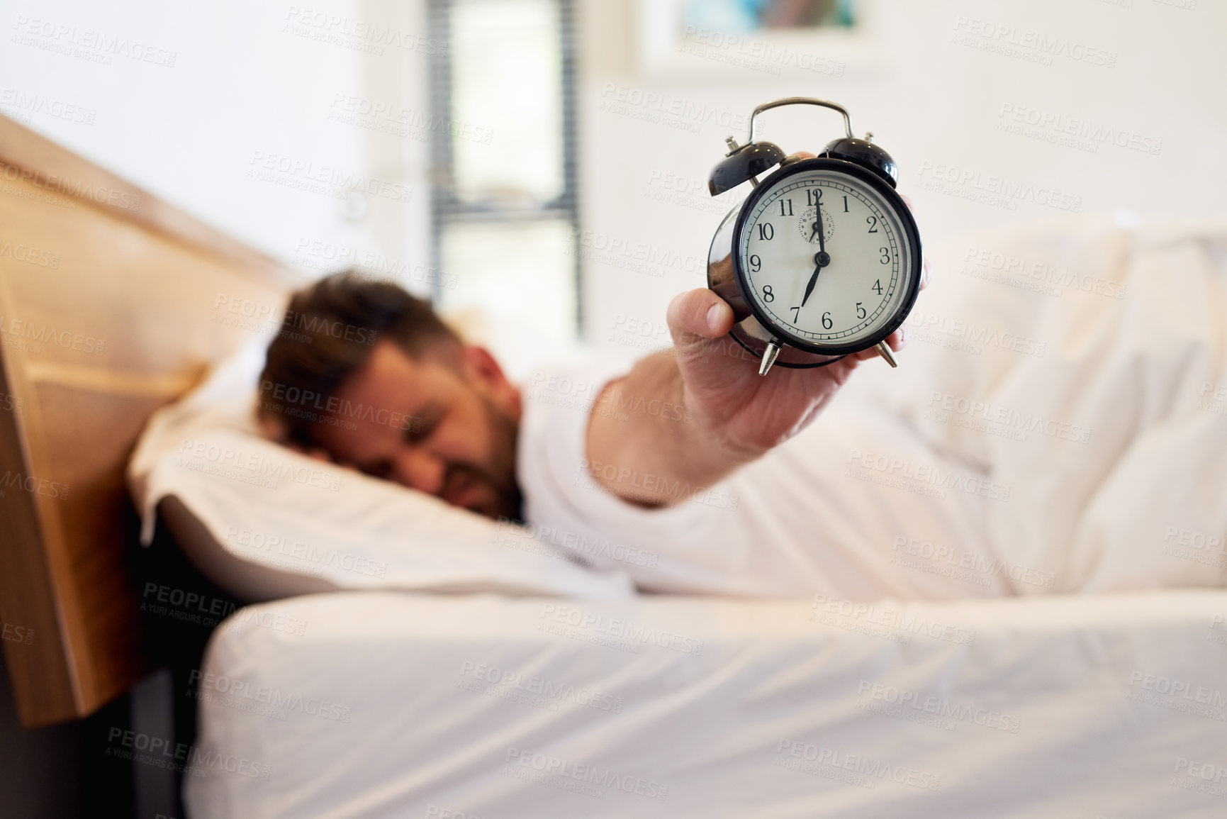 Buy stock photo Cropped shot of a young man holding up an alarm clock while waking up from bed at home
