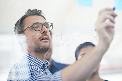 Buy stock photo Cropped shot of a handsome businessman making notes on a glass wall while his colleagues look on