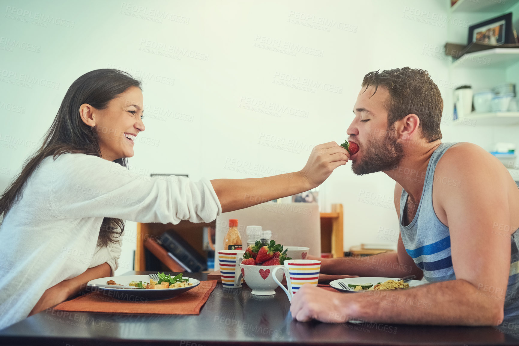 Buy stock photo Shot of a happy young woman feeding a strawberry to her boyfriend over the breakfast table