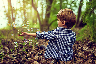 Buy stock photo Shot of a little boy playing with bubbles while sitting alone in the forest