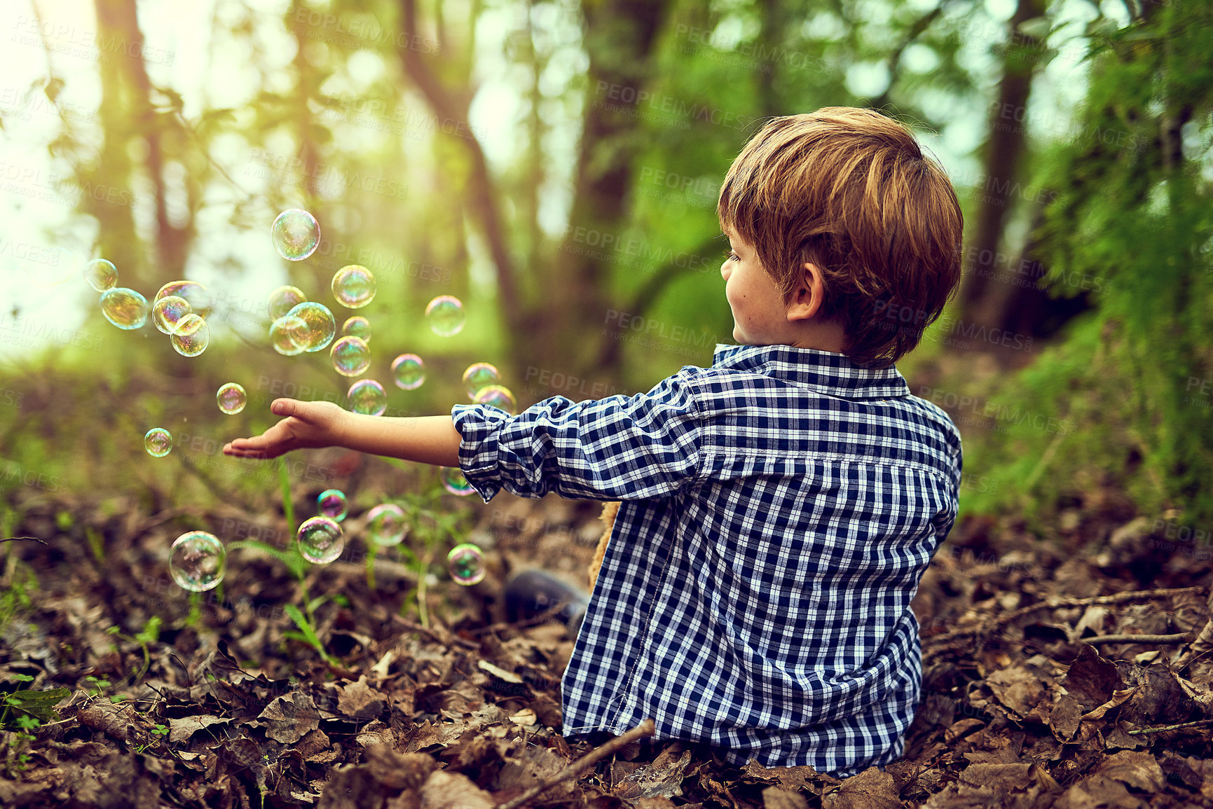 Buy stock photo Shot of a little boy playing with bubbles while sitting alone in the forest