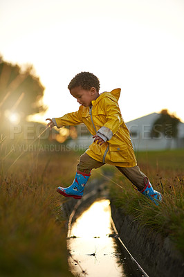 Buy stock photo Winter, boy and jump with raincoat by water for playful, adventure and holiday fun at sunset. Nature, black kid and happy by drainage system in grass for cold weather, freedom and child development