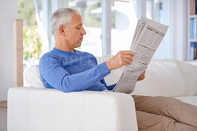 Buy stock photo Shot of a mature man sitting on his living room sofa reading a newspaper