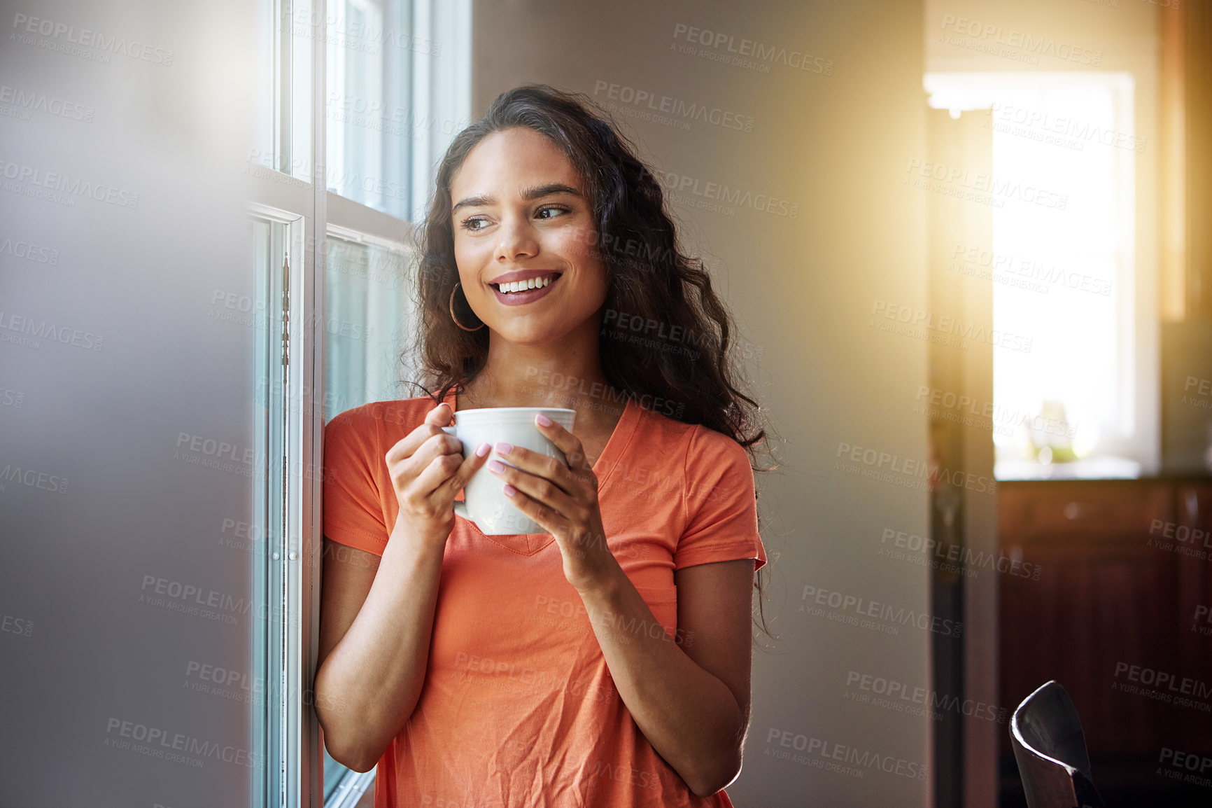 Buy stock photo Shot of a smiling young woman looking through a window while drinking a coffee at home