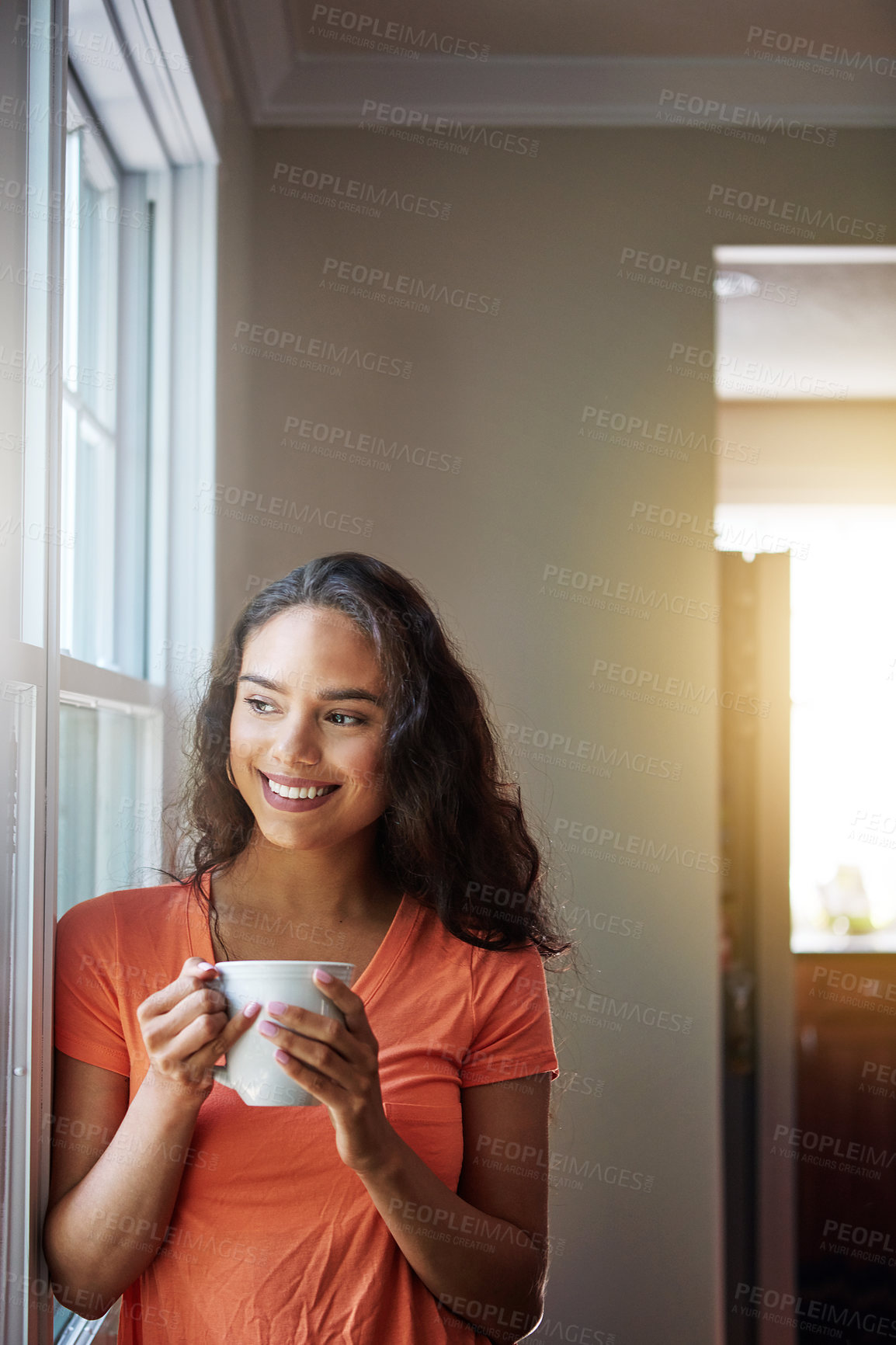 Buy stock photo Shot of a smiling young woman looking through a window while drinking a coffee at home