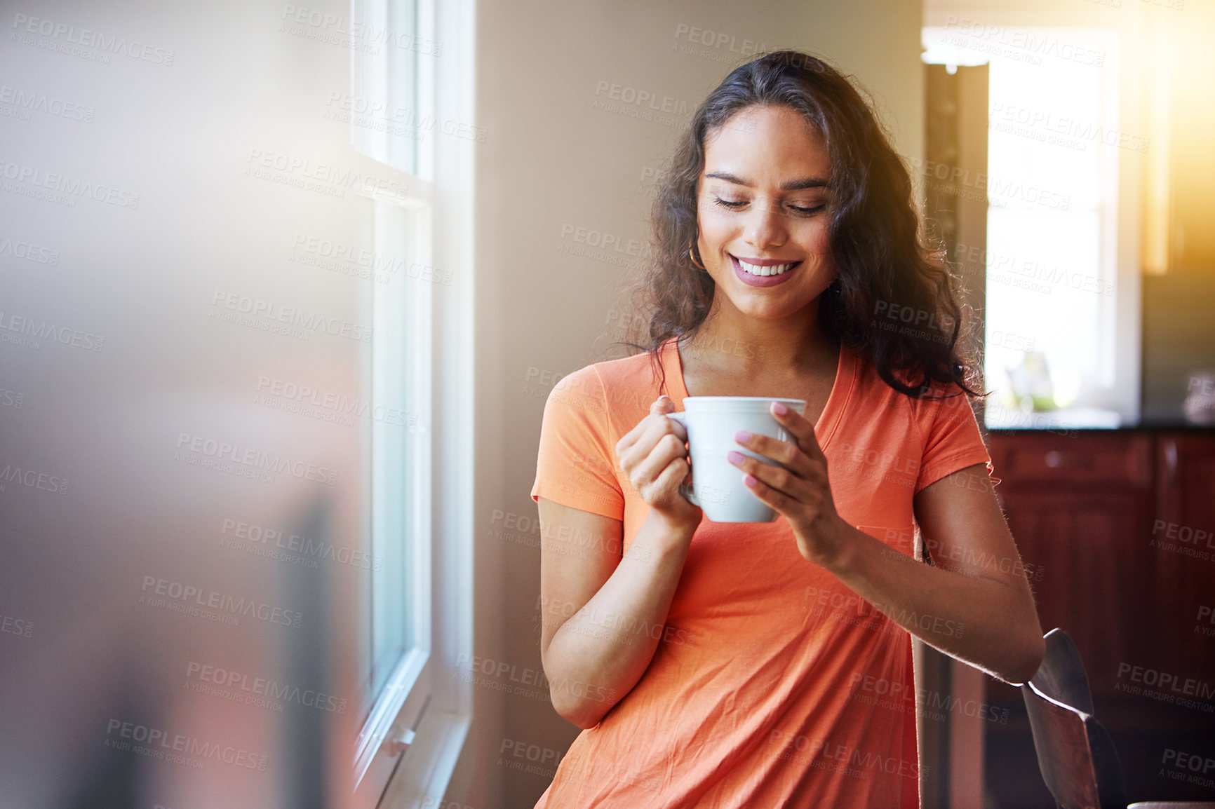 Buy stock photo Shot of a smiling young woman looking through a window while drinking a coffee at home