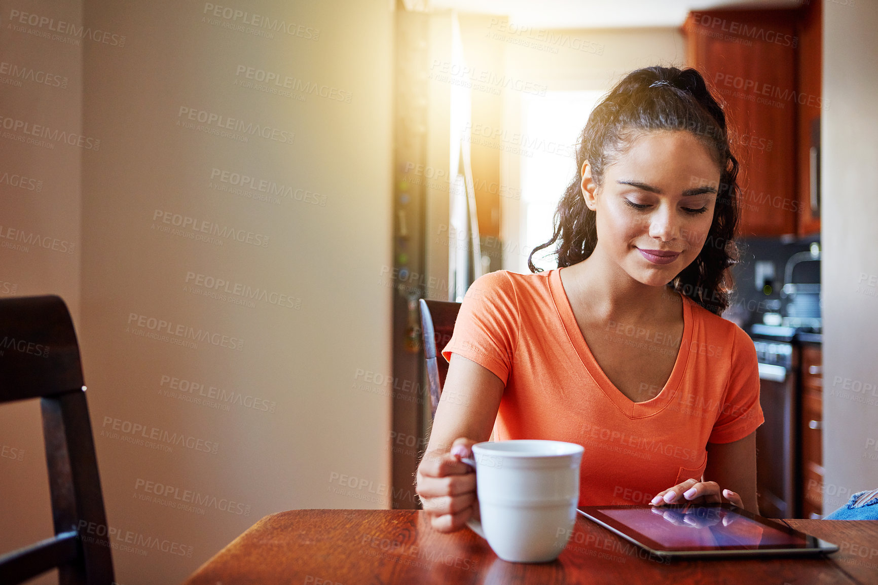 Buy stock photo Shot of a smiling young woman sitting at her dining table at home drinking coffee and using a digital tablet