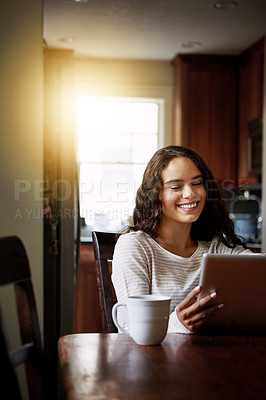 Buy stock photo Shot of a smiling young woman sitting at her dining table at home drinking coffee and using a digital tablet