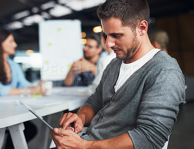 Buy stock photo Shot of a young man sitting at a table in an office using a digital tablet with colleagues working in the background