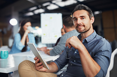 Buy stock photo Portrait of a man sitting at a table in an office using a digital tablet with colleagues working in the background