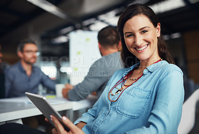 Buy stock photo Portrait of a woman sitting at a table in an office using a digital tablet with colleagues working in the background