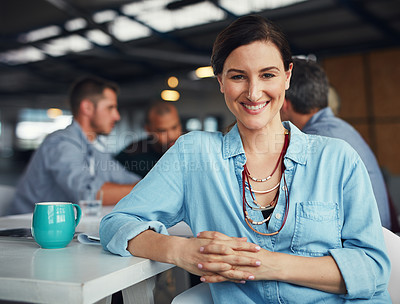 Buy stock photo Portrait of a smiling woman sitting at a table in an office with colleagues working in the background