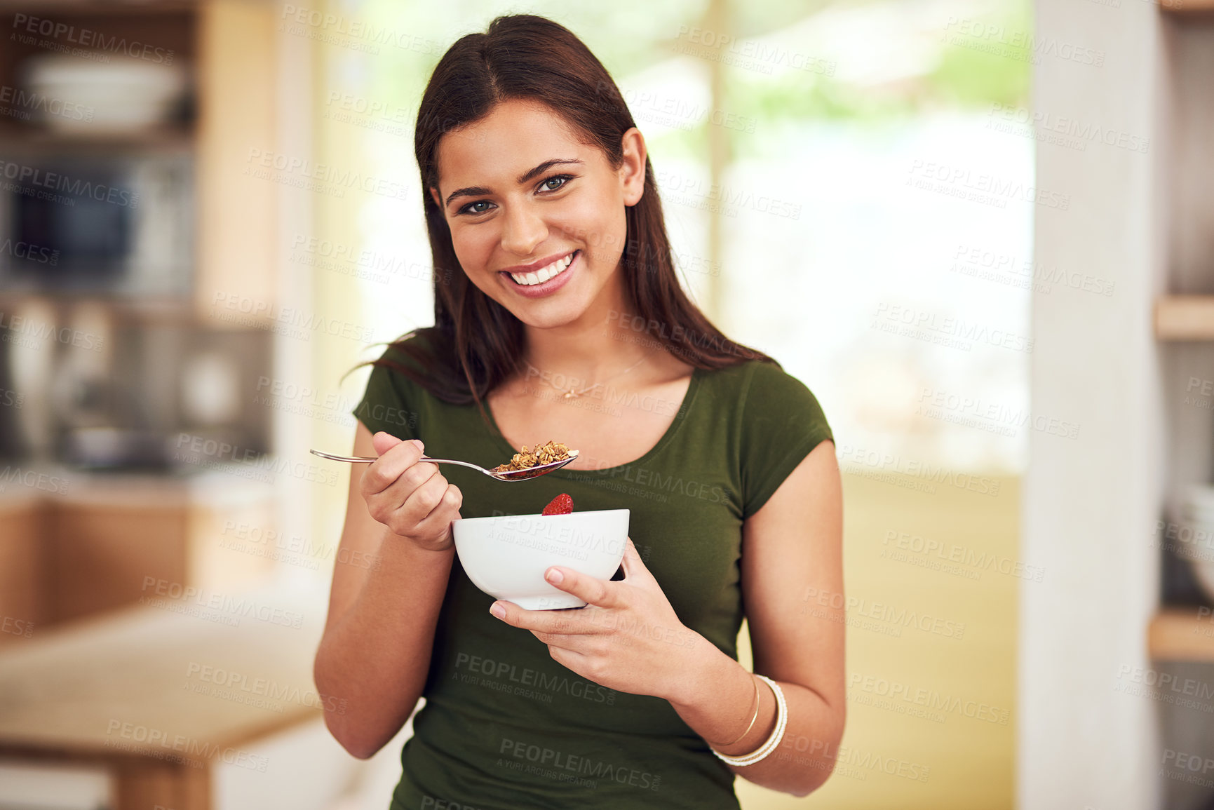 Buy stock photo Portrait of a happy young woman eating a bowl of muesli while standing in her kitchen