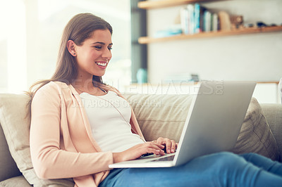 Buy stock photo Shot of a smiling young woman using a laptop while relaxing on the sofa at home