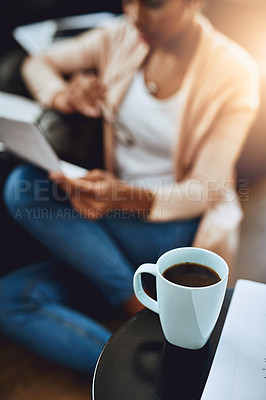 Buy stock photo Cropped shot of a cup of coffee on a table with a woman reading a document in the background at home