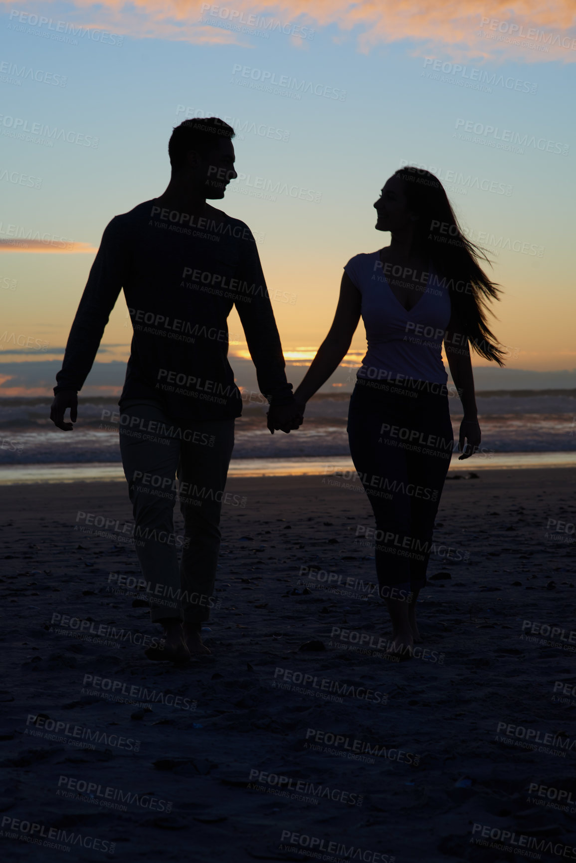 Buy stock photo Silhouette shot of a young couple walking on the beach