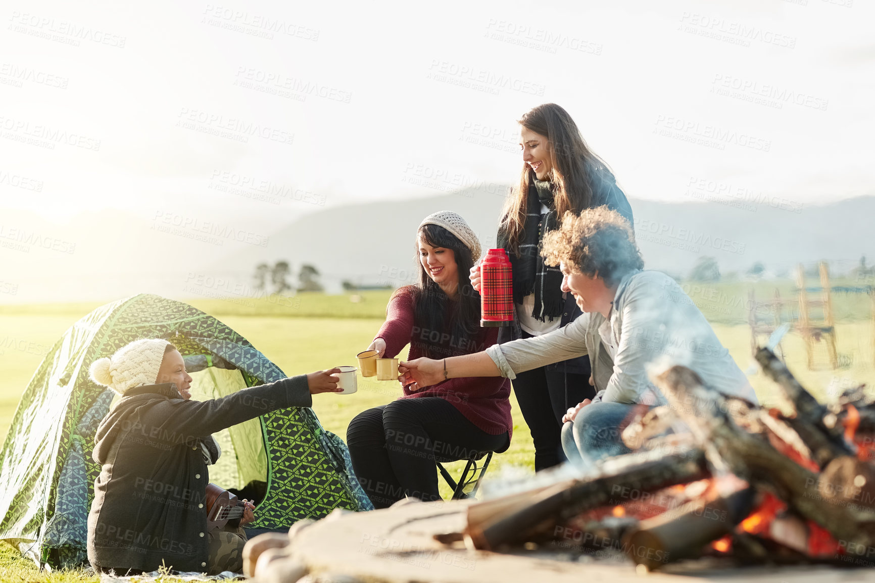 Buy stock photo Shot of a group of young friends enjoying coffee by the fire while on a camping trip