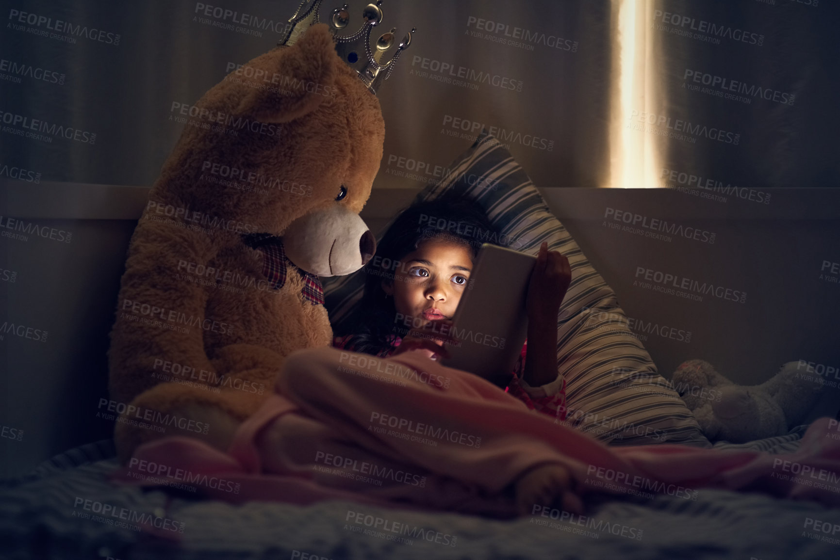 Buy stock photo Shot of a little girl using a digital tablet while lying in bed with her teddy at night