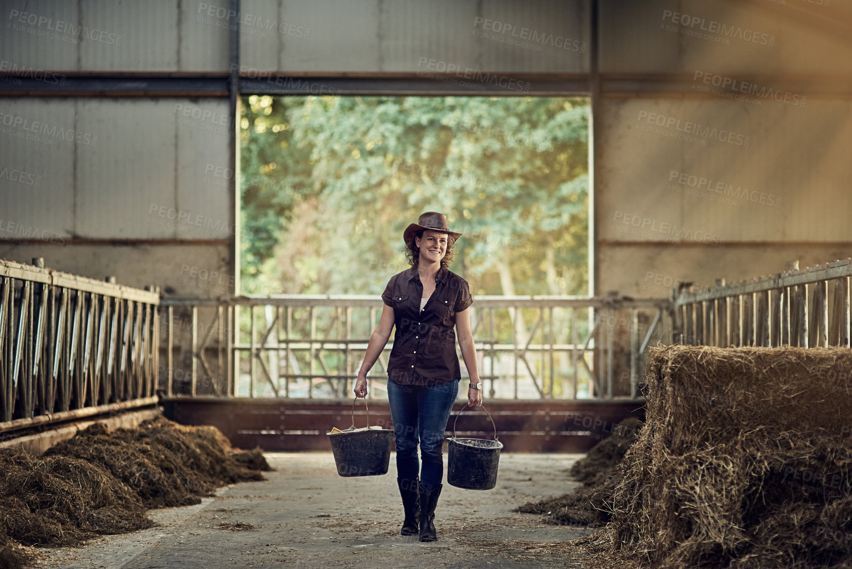 Buy stock photo Shot of a happy female farmer carrying buckets while walking out of a barn