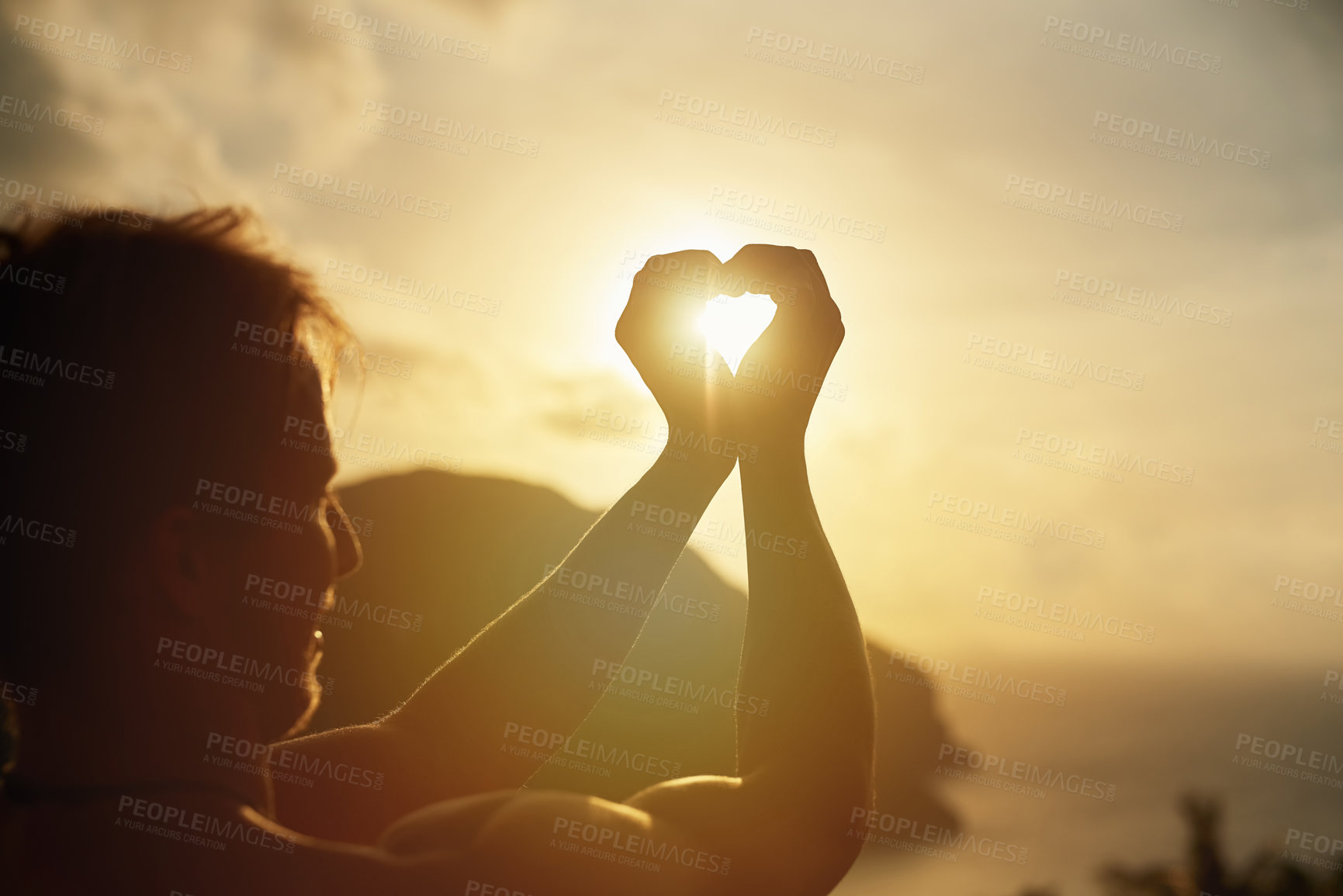 Buy stock photo Shot of a young man making a heart shape with his hands around the sun while on holiday