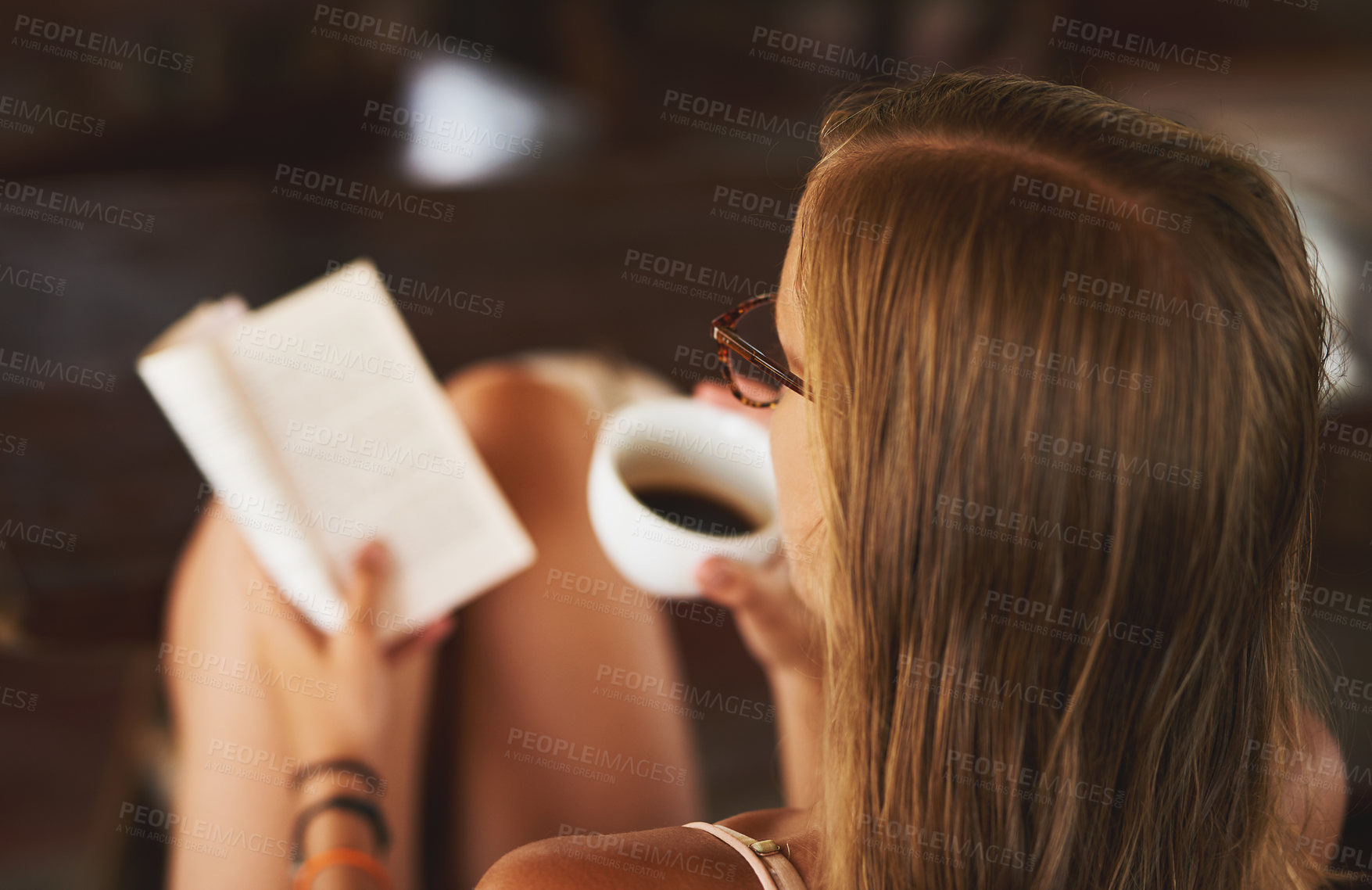 Buy stock photo Rear view shot of a young woman reading a book while drinking a cup of coffee