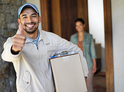Buy stock photo Portrait of a smiling young courier giving a thumbs up and carrying a package with a customer standing at her front door in the background