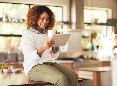 Buy stock photo Shot of a happy young woman using a digital tablet and drinking coffee in the kitchen at home