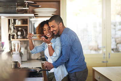 Buy stock photo Shot of a young woman giving her husband a taste of the food that she’s preparing at home