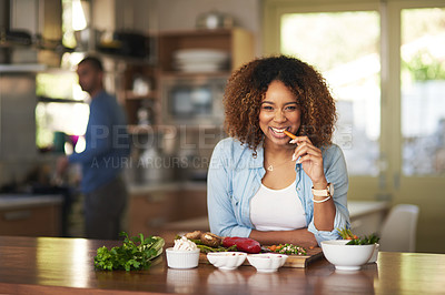 Buy stock photo Portrait of a young woman eating a carrot while preparing a healthy meal with her husband in the background