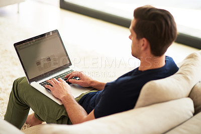 Buy stock photo High angle shot of a young man using his laptop while sitting on the sofa at home