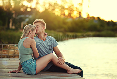 Buy stock photo Shot of an affectionate young couple sitting together at a lake outside