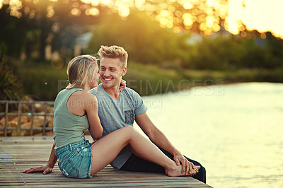 Buy stock photo Shot of an affectionate young couple sitting together at a lake outside
