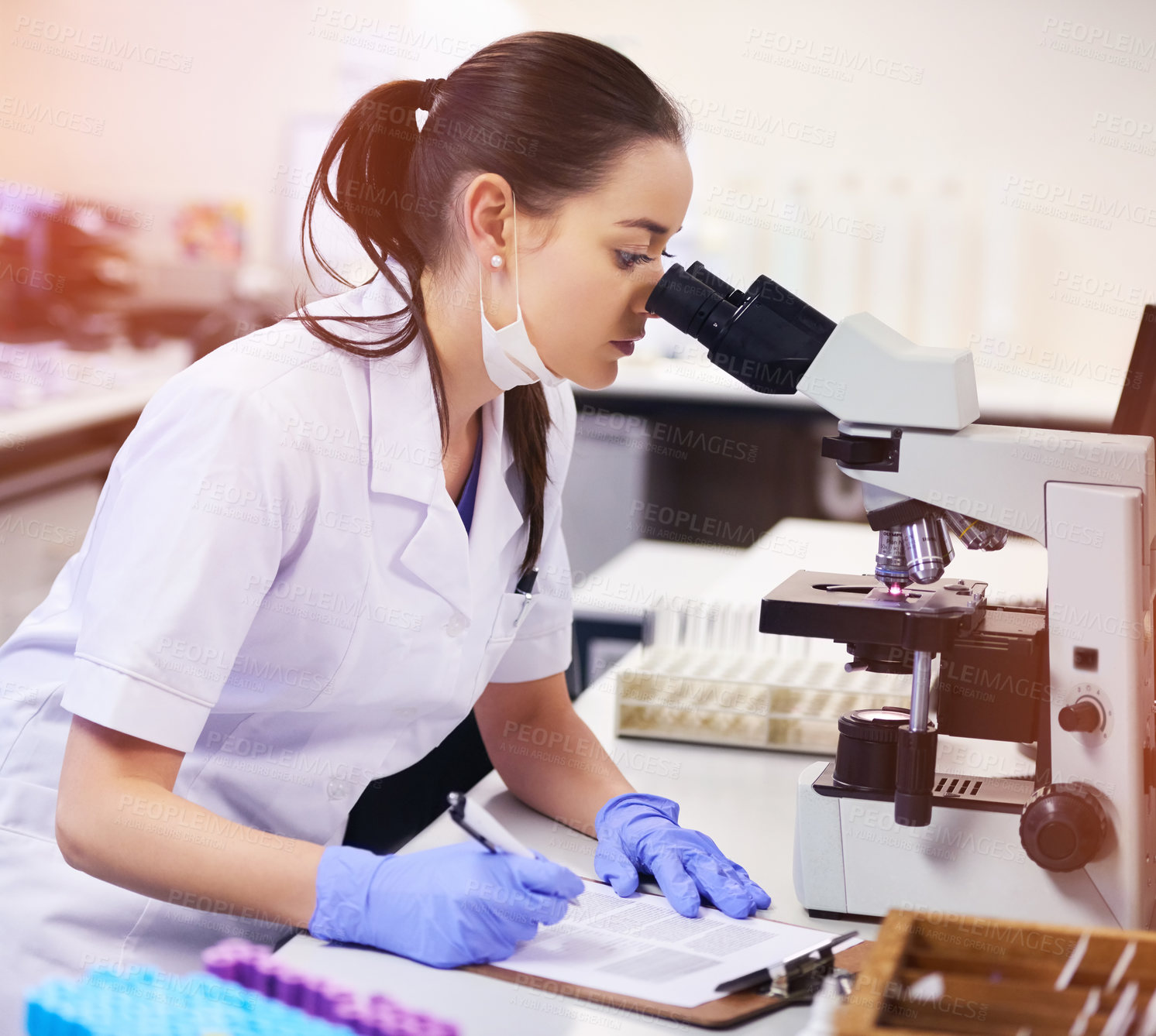 Buy stock photo Shot of a young scientist taking notes while using a microscope in a laboratory