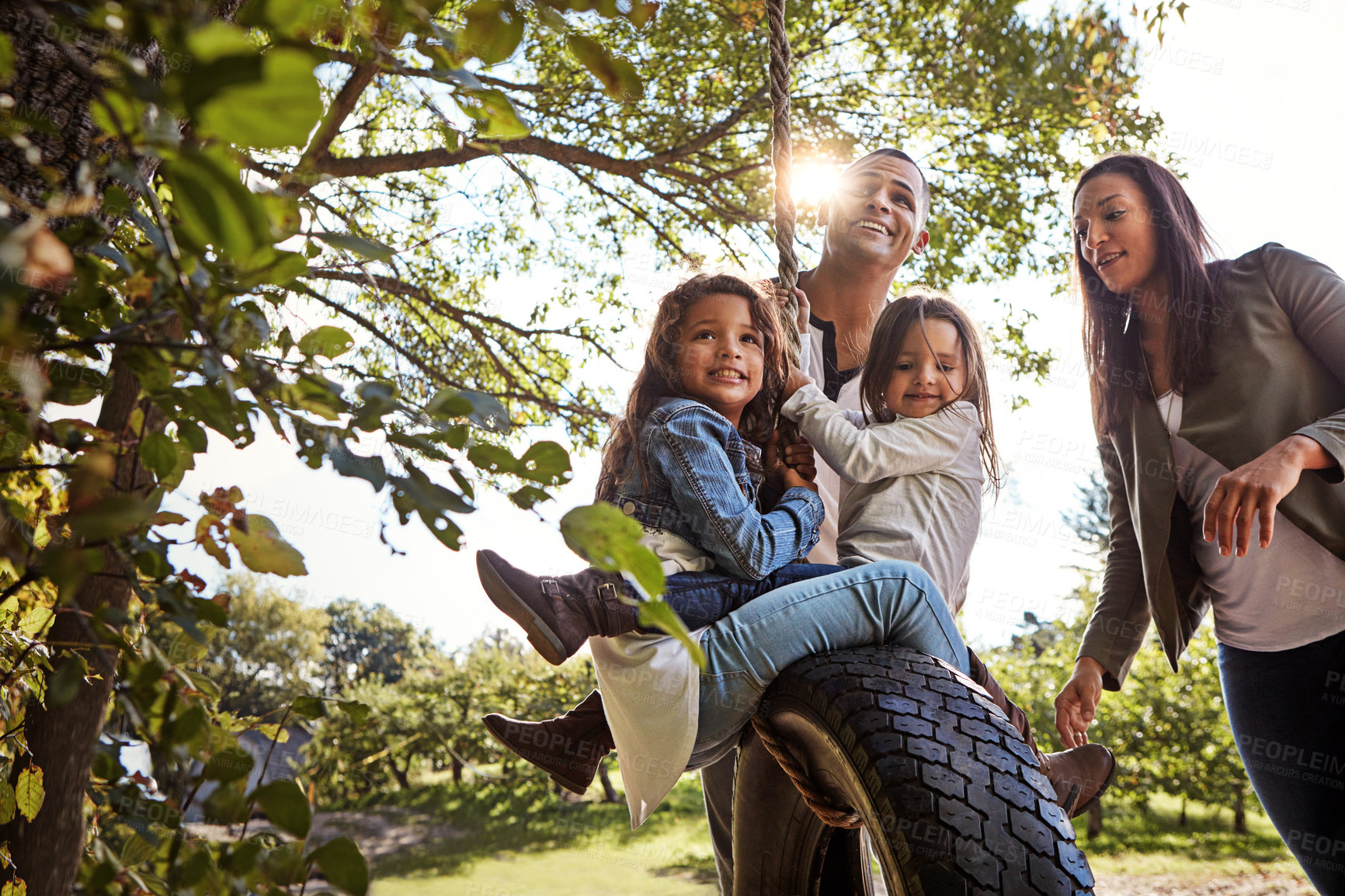 Buy stock photo Shot of a happy mother and  father pushing their daughters on a tyre swing