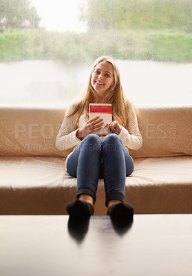 Buy stock photo Portrait of an attractive young woman holding a book while sitting on the sofa at home