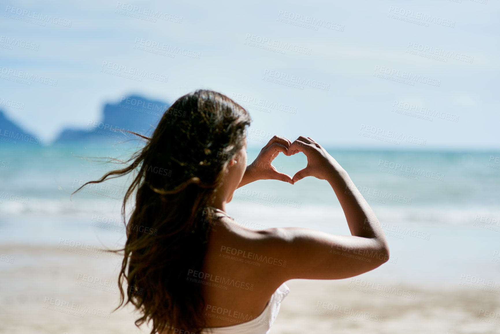 Buy stock photo Shot of a young woman shaping a heart while looking at the scenery at the beach