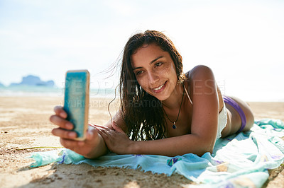 Buy stock photo Shot of a young woman taking a selfie while lying on the beach