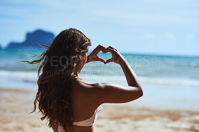 Buy stock photo Shot of a young woman shaping a heart while looking at the scenery at the beach