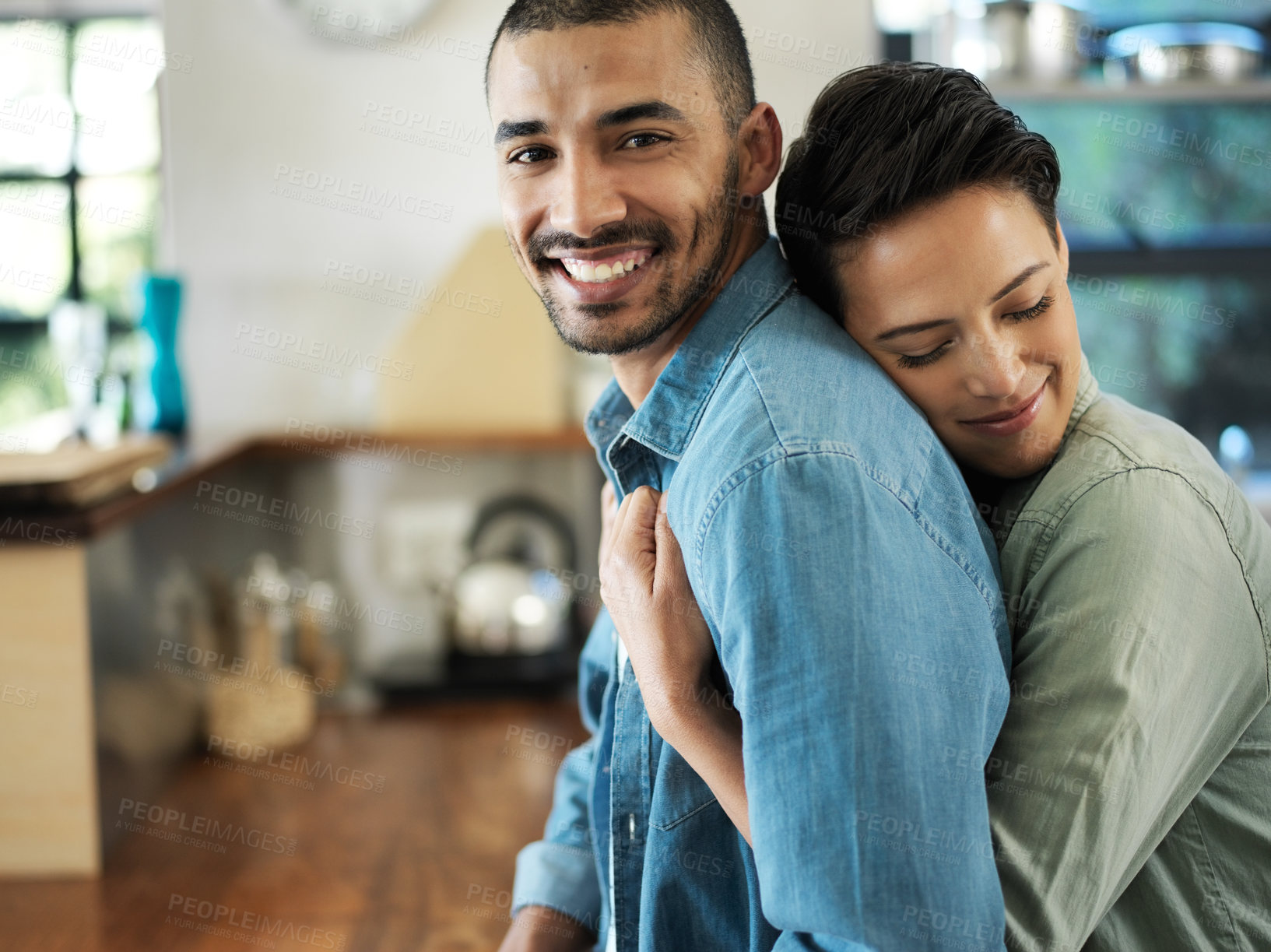 Buy stock photo Shot of a young woman embracing her husband from behind while standing in their kitchen