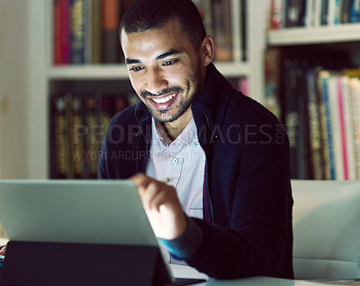 Buy stock photo Shot of a smiling young man working on a digital tablet in his home office in the early evening