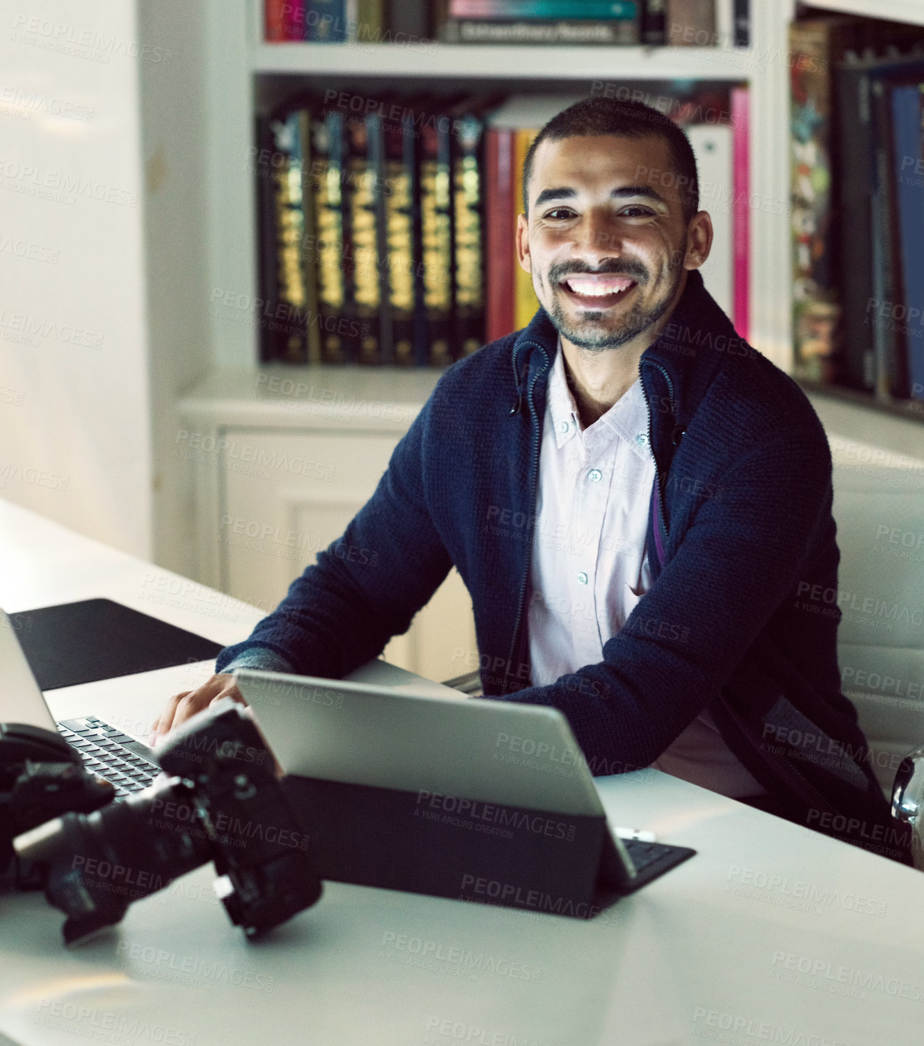 Buy stock photo Portrait of a smiling young photographer working on a digital tablet and laptop in his home office in the early evening