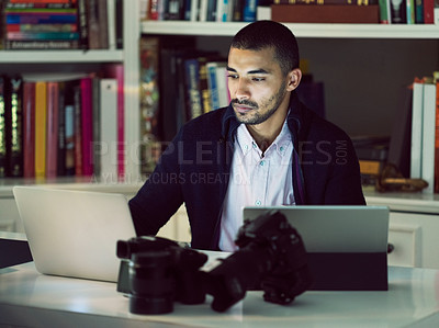 Buy stock photo Shot of a focused young photographer working on a digital tablet and laptop in his home office in the early evening