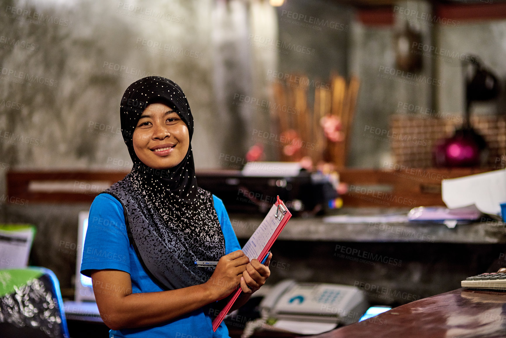 Buy stock photo Portrait of a smiling muslim thai woman standing behind a hotel reception desk holding a clipboard