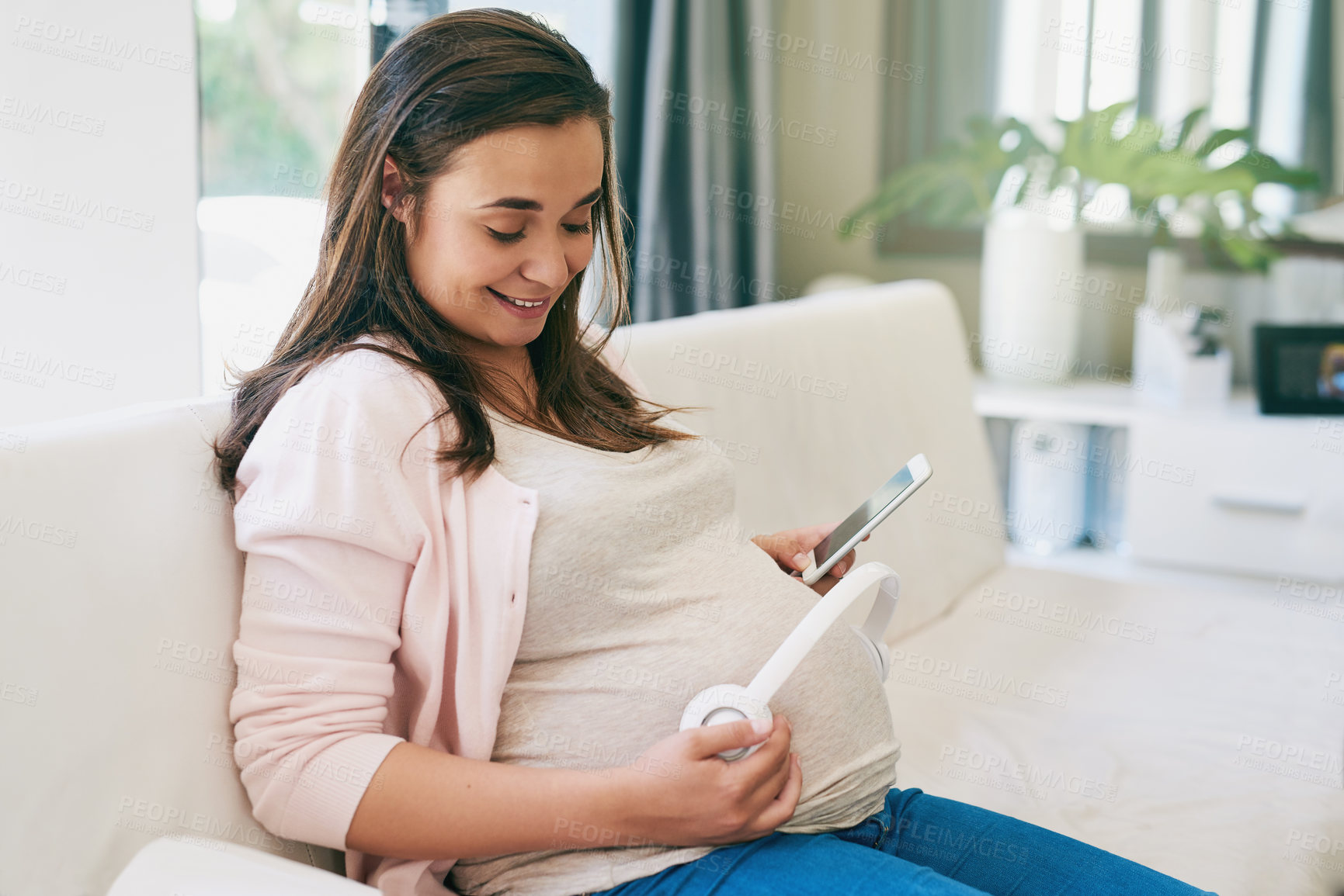 Buy stock photo Relax, phone and pregnant woman on couch with headphones on stomach for child development in home. Pregnancy, growth and mom on sofa with smartphone, sound and app for music streaming in living room