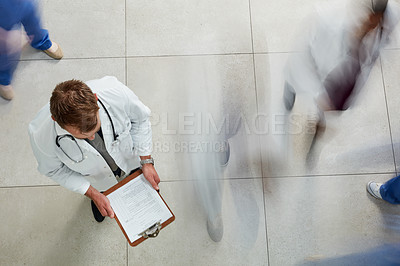 Buy stock photo High angle shot of a male doctor looking at a patient's file in a busy hospital