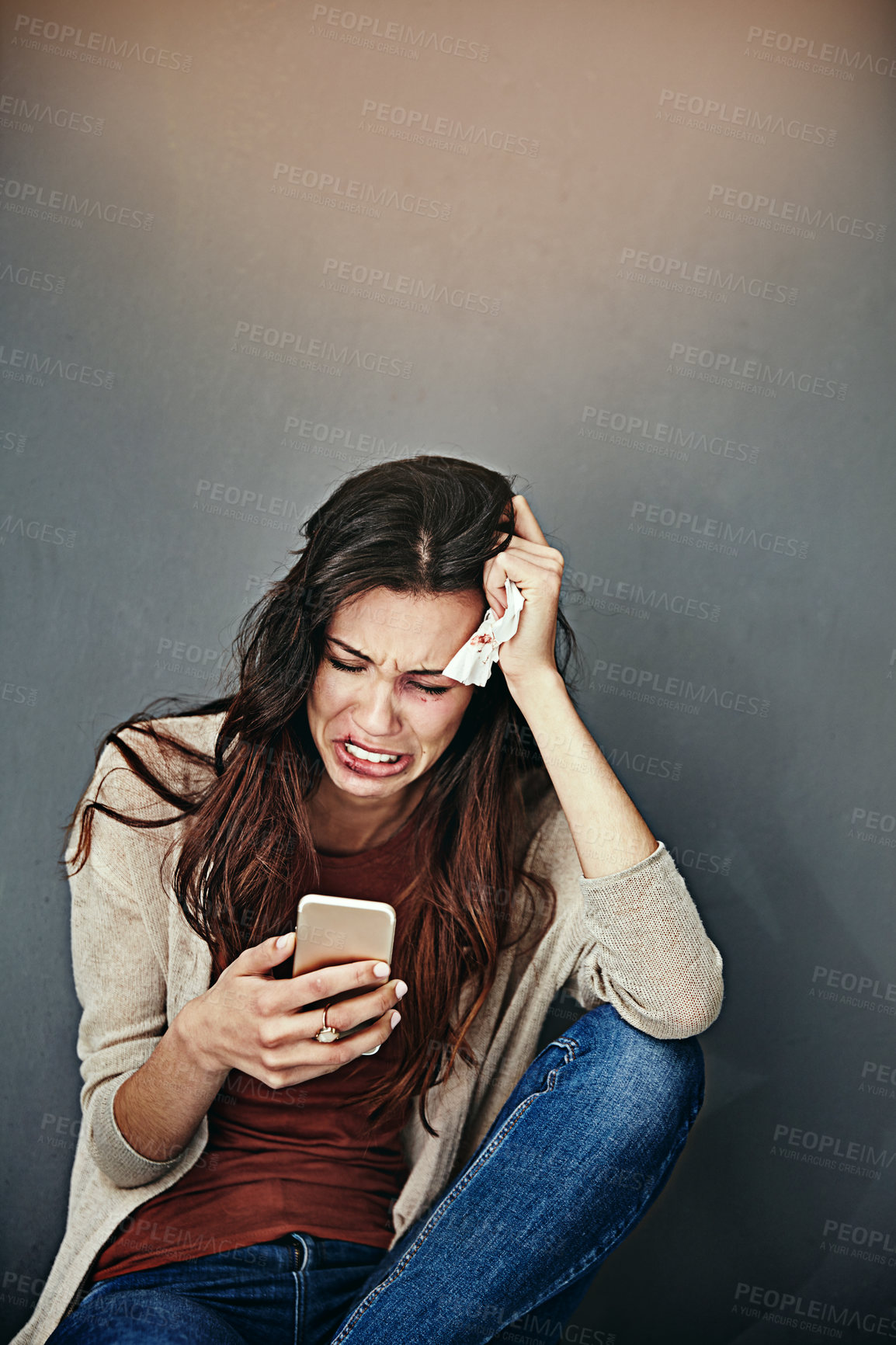 Buy stock photo Shot of a beaten and bruised young woman crying while looking at her cellphone