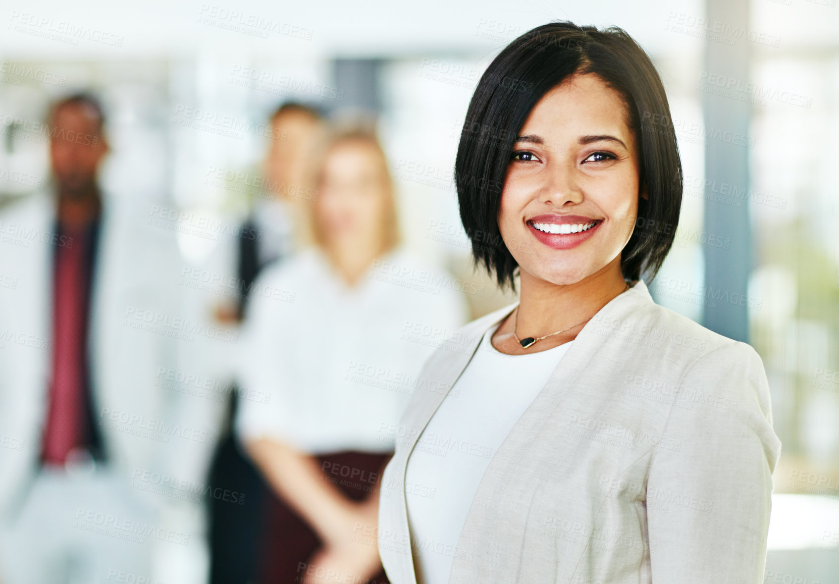 Buy stock photo Young, smart and smiling businesswoman with a diverse team at work ready to achieve success. Confident female professional executive standing in a modern office with her colleagues in the background