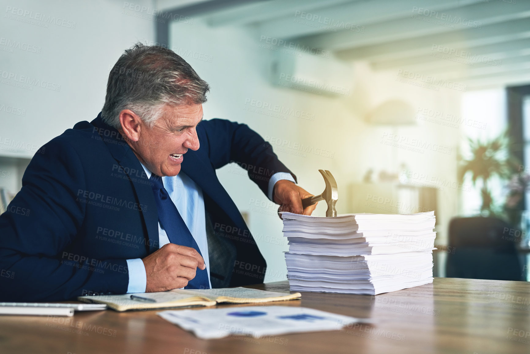 Buy stock photo Shot of a frustrated businessman hitting a pile of paperwork with a hammer