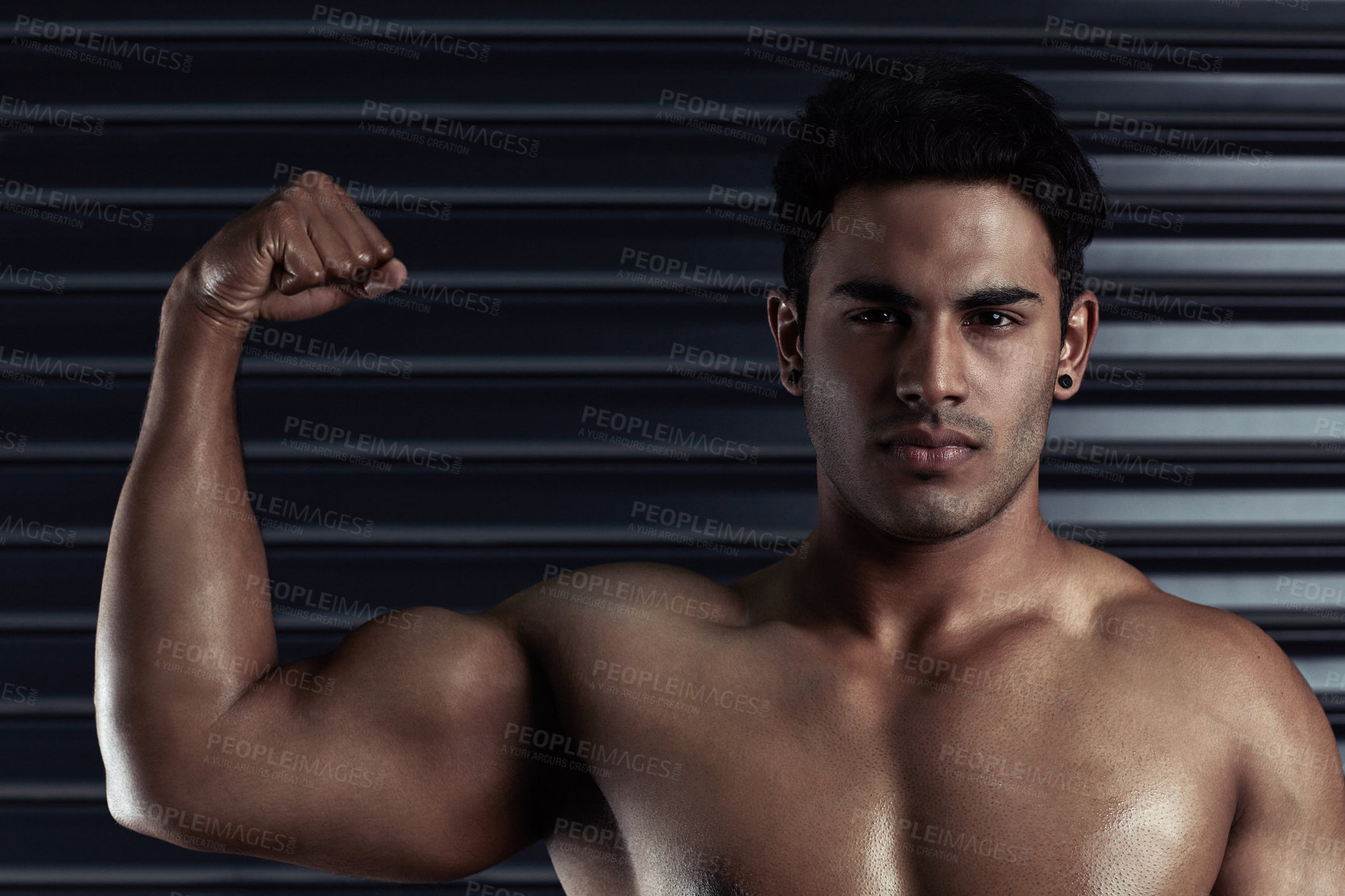 Buy stock photo Cropped portrait of an athletic young man flexing his bicep against a dark background